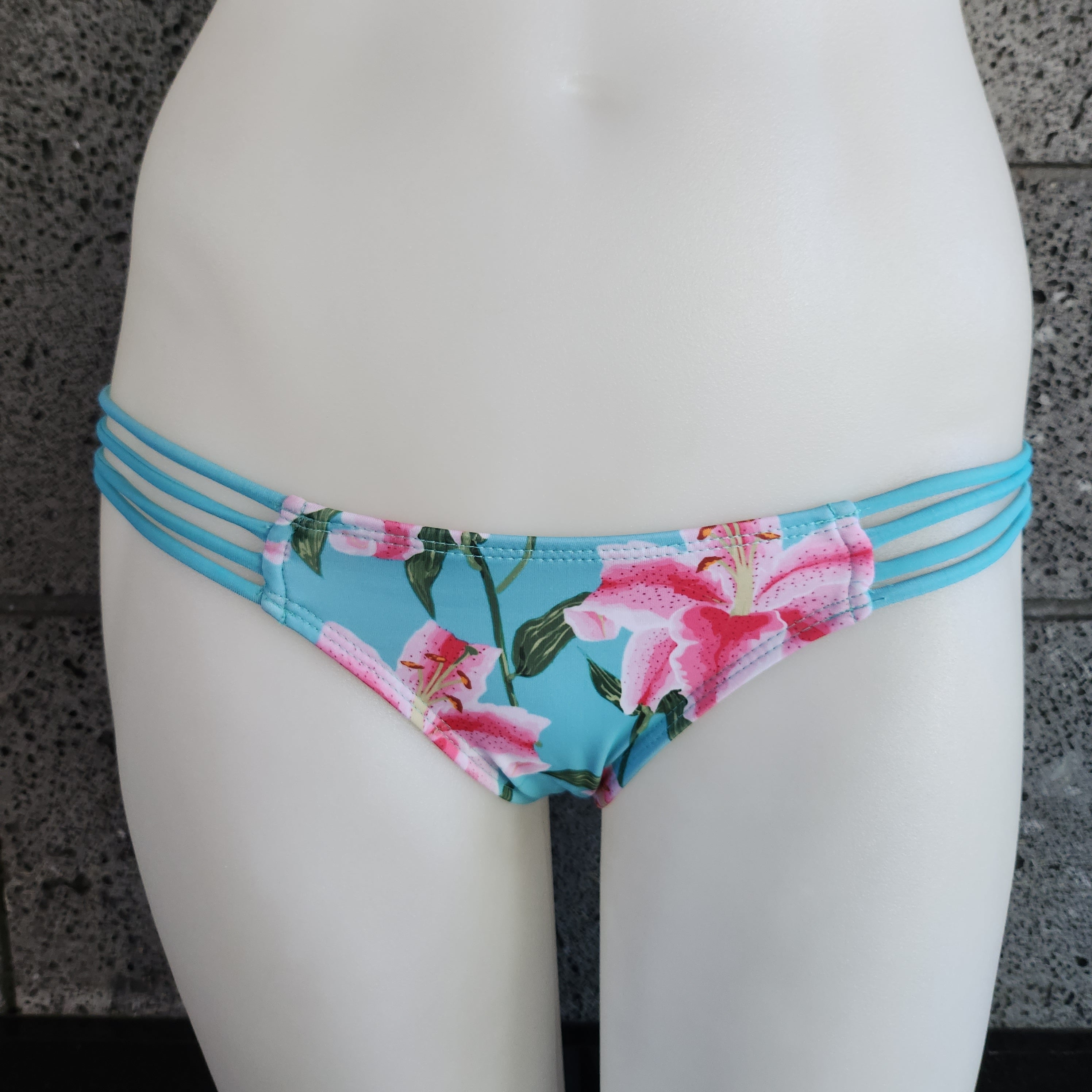 Floral bikini bottom with strappy sides on a mannequin against a gray concrete wall.