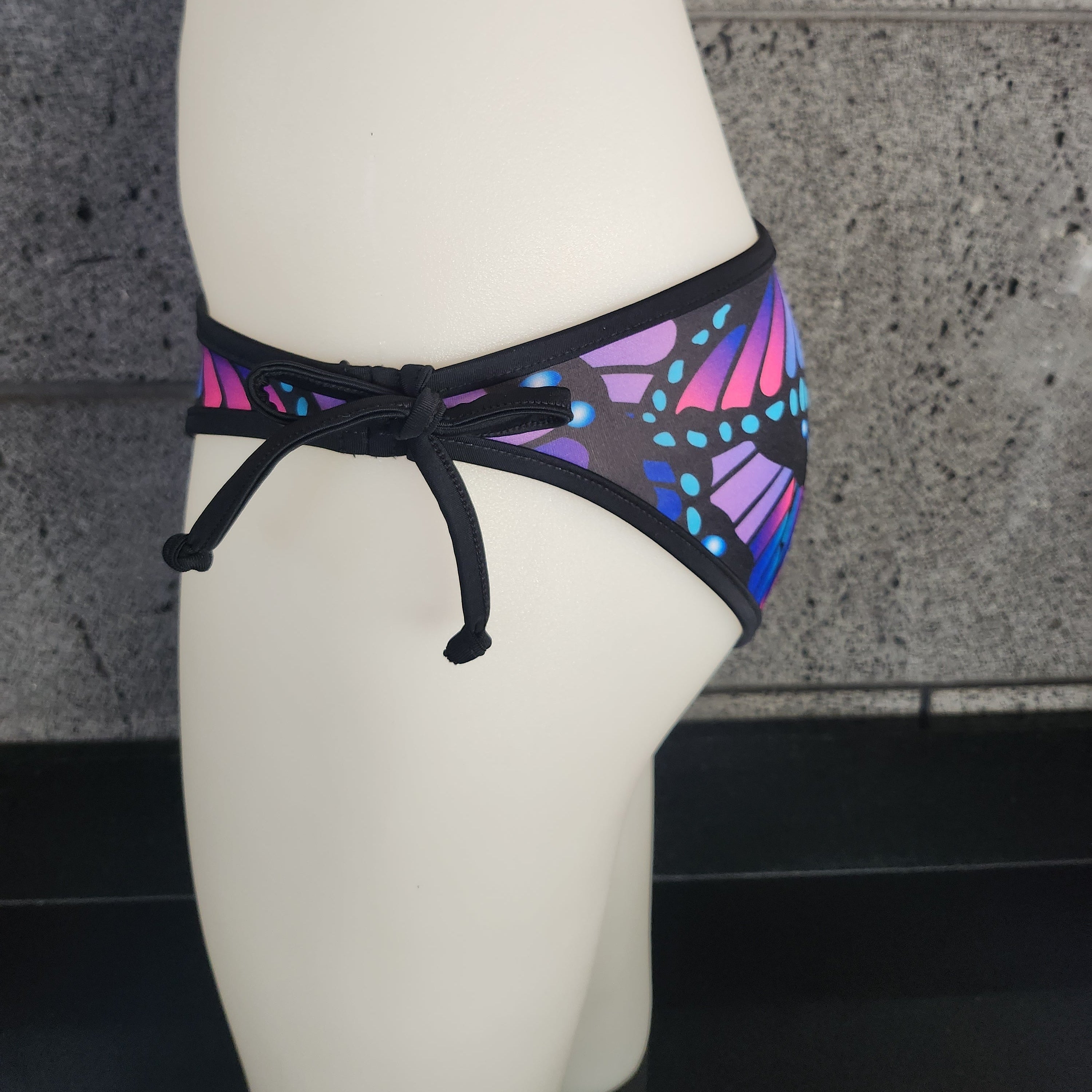 Multicolored butterfly-patterned bikini bottom on a mannequin against a gray textured wall.