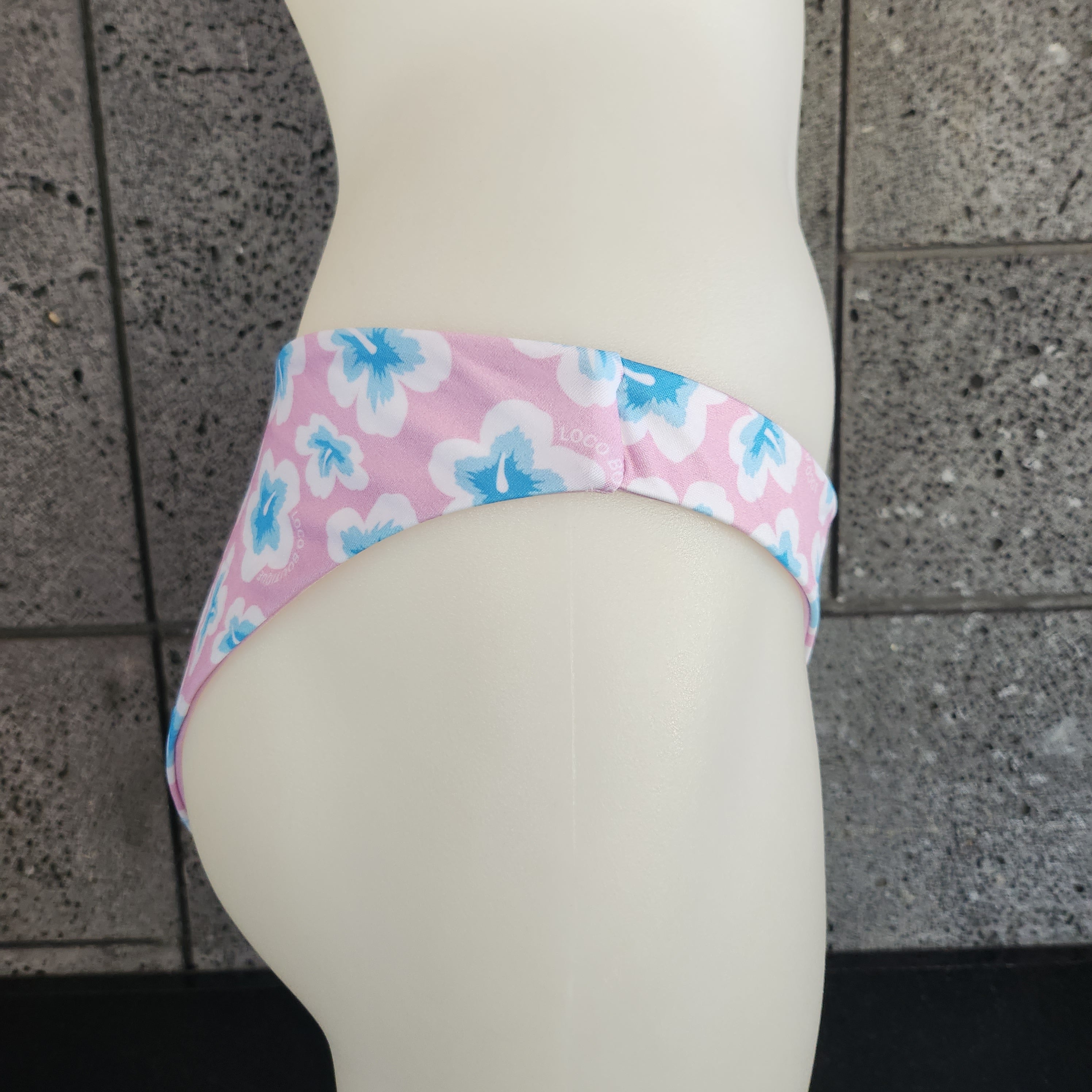 Blue bikini bottom with pink floral pattern on a mannequin against a concrete wall.