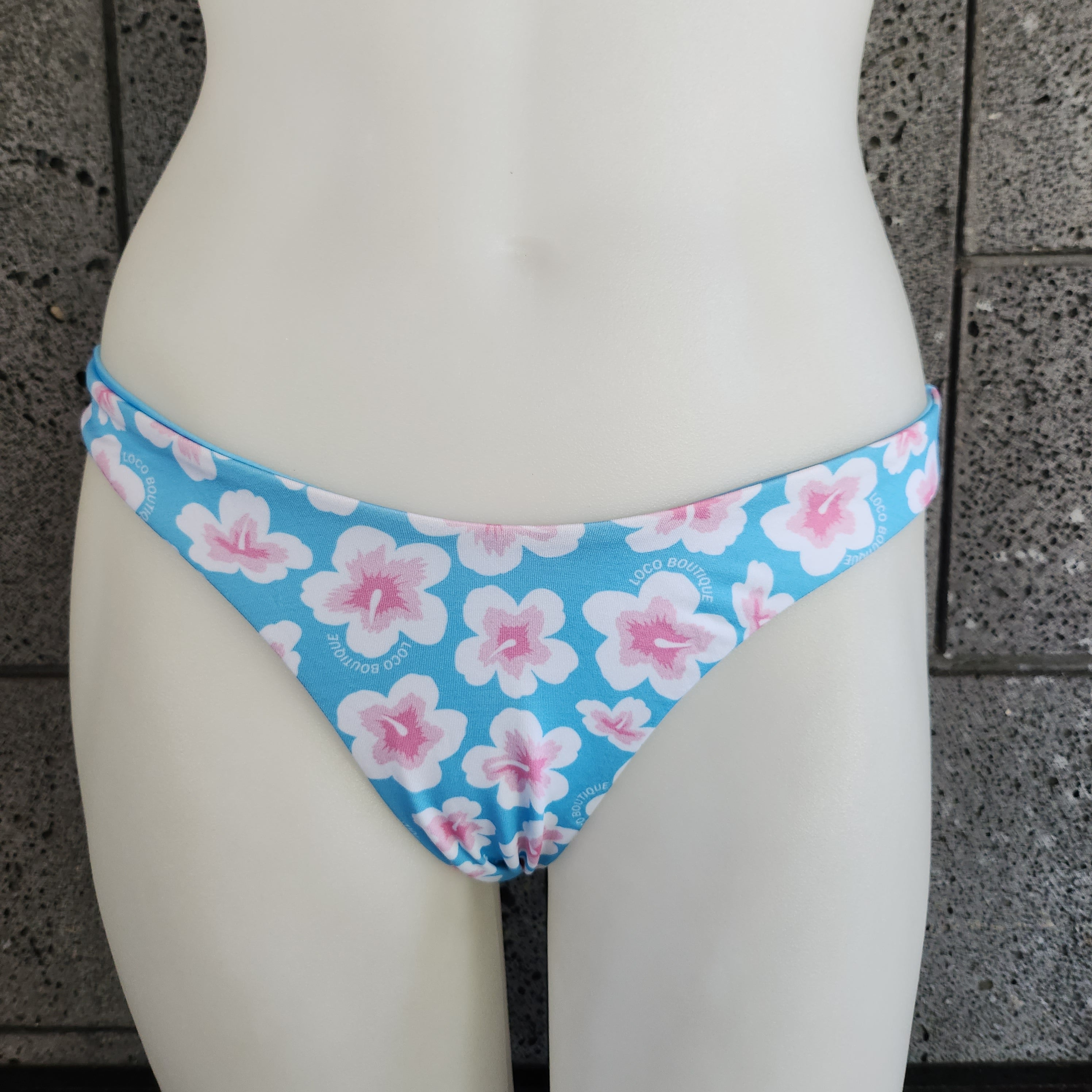 Blue bikini bottom with pink floral pattern on a mannequin against a concrete wall.